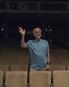 Erica Hong/Courier Theatre instructor and director, Whitney Rydbeck waves "goodbye" in the audience seating of Sexson Auditorium on Wednesday, July 08, 2015 at PCC. Rydbeck has retired after four decades at PCC but cheerfully returned to be photographed on the other side of the stage, as posters of his final directorial performance, Spoon River still hang on the walls.